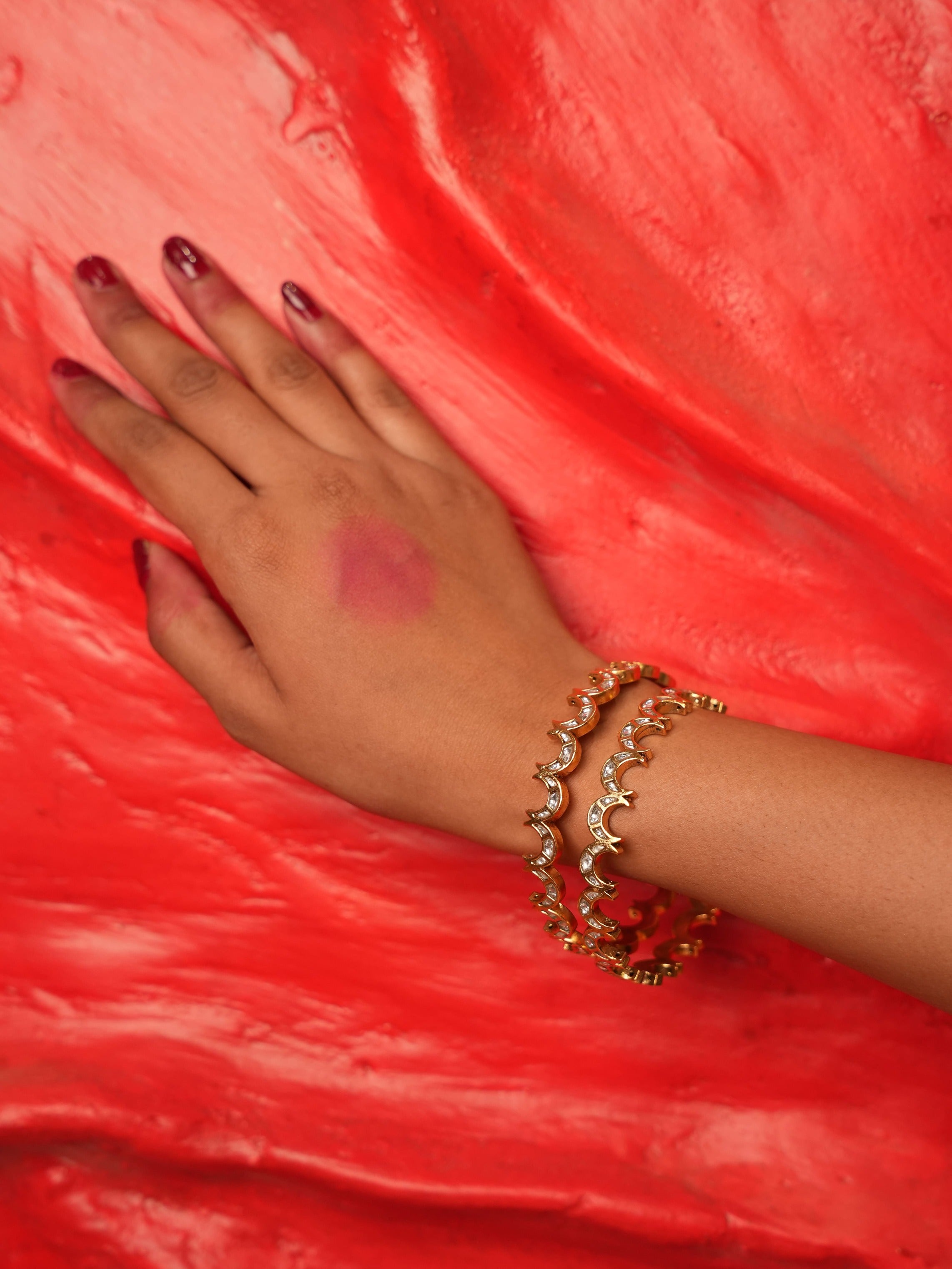 A close-up of a person's hand and wrist wearing three gold crescent moon beaded bracelets, set against a vibrant red textured background.